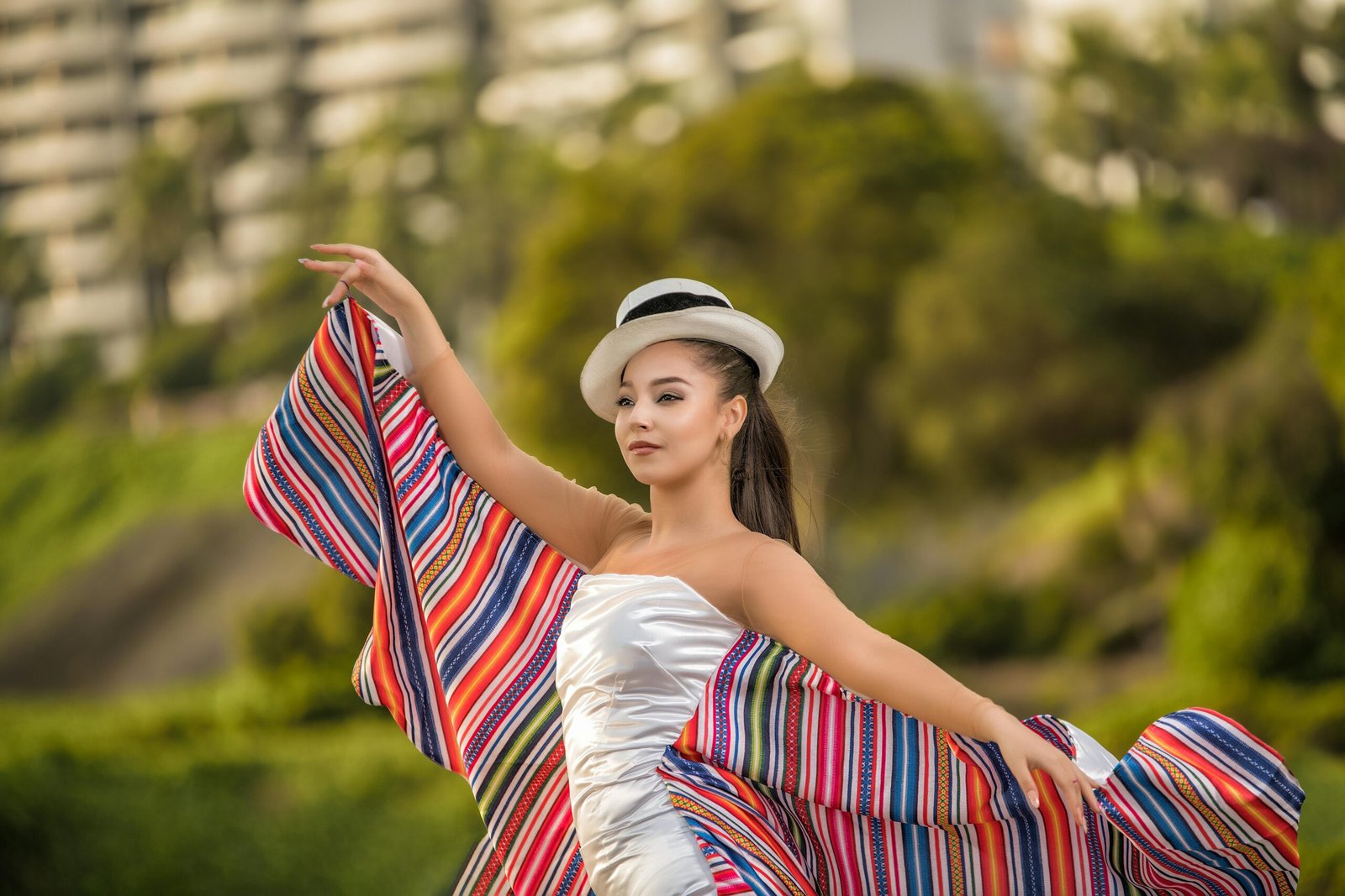 Elegant woman posing in a striped dress and hat in an outdoor setting with lush greenery.